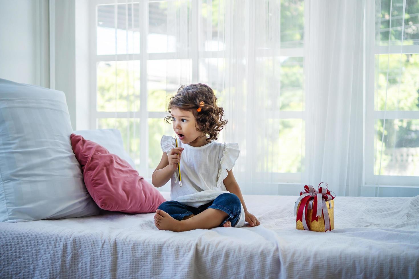 toma felicitaciones por tu cumpleaños. una niña pequeña con rulos se sienta en la cama del dormitorio jugando con un lápiz. preparación de la fiesta. niño divertido con un lápiz. foto