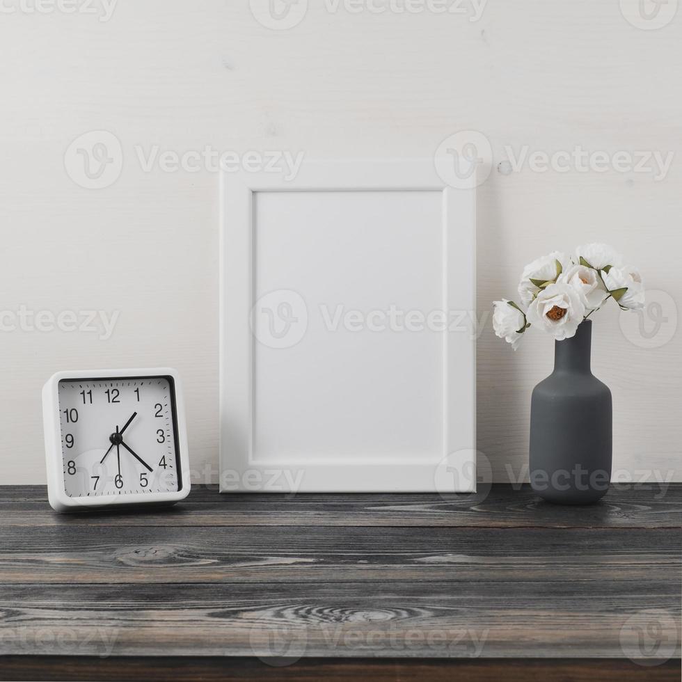 White frame, flower in vaze, clock on dark gray wooden table against the white wall with copy space. Mock up. photo