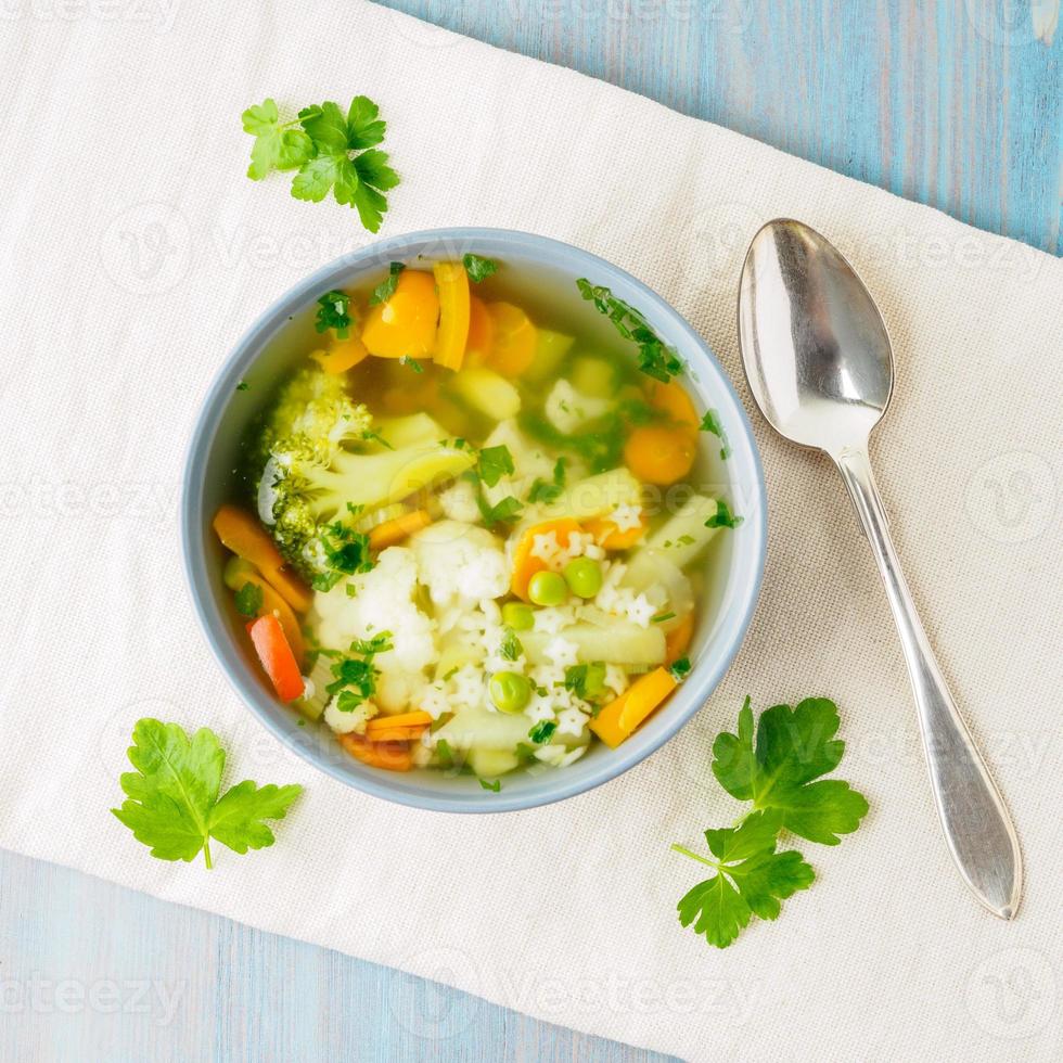 Bright spring vegetable dietary vegetarian soup with cauliflower, broccoli, pepper, carrot, green peas, pasta, parsley. Top view, blue background. photo