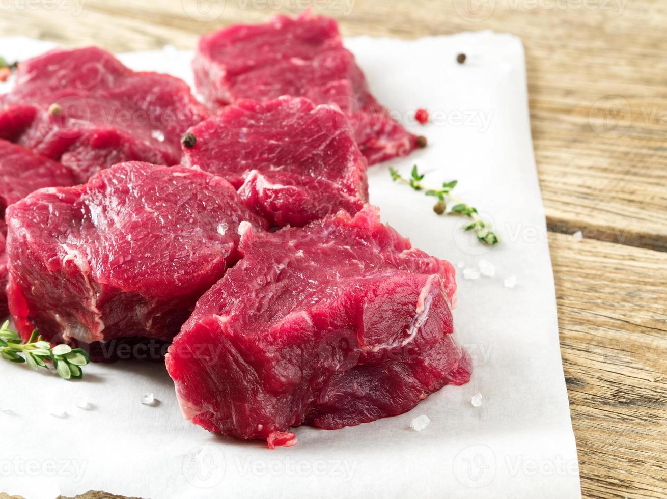 Pieces of raw meat. Raw beef with spices on white parchment paper on wooden rough rustic background, side view, close-up photo