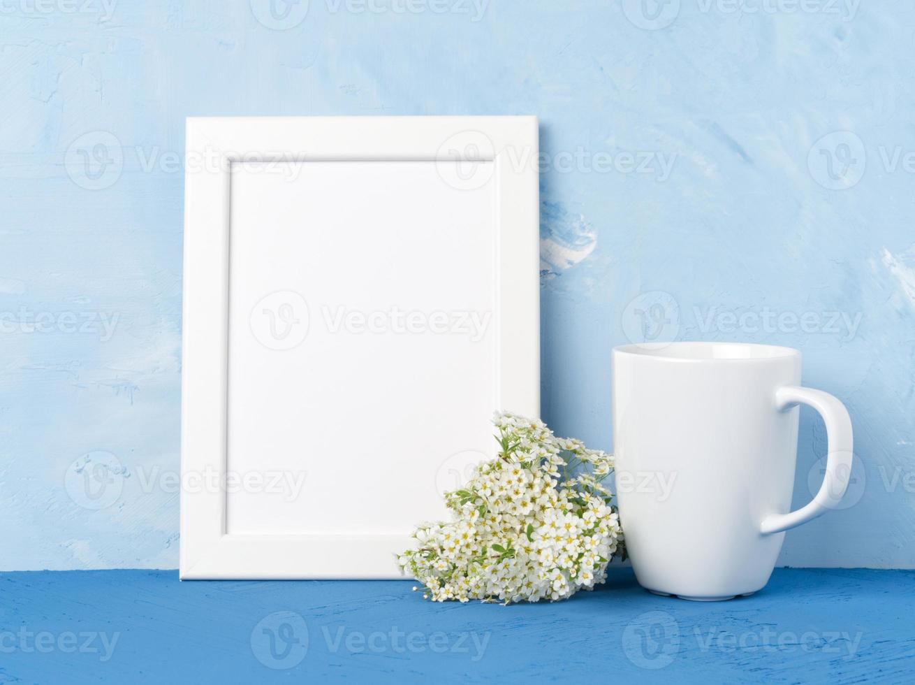 White mug with tea or coffee, frame, flower bouquet on blue table opposite blue concrete wall. Mock up with empty space photo