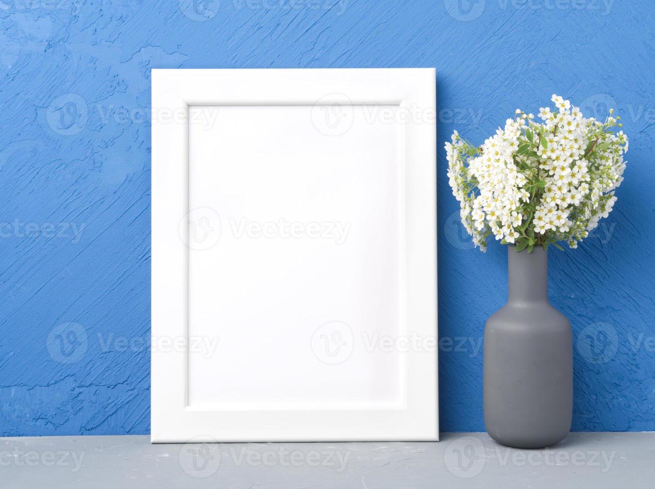 Blank white frame, flower in vaze on gray table against dark blue concrete wall photo