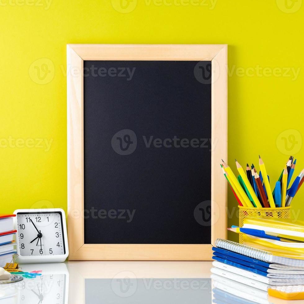 pizarra, libros de texto, reloj y útiles escolares en la mesa junto a la pared amarilla. vista lateral, espacio para texto. concepto de regreso a la escuela. foto