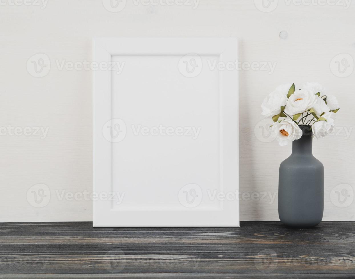 White frame, flower in vase, clock on dark grey wooden table against white wall with copy space photo