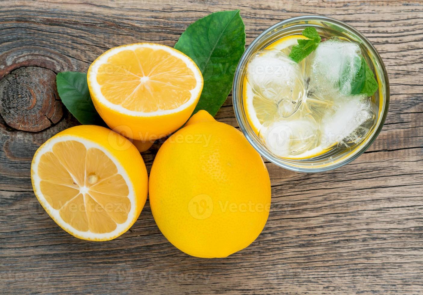 Lemonade in a glass, a lemon half, fresh leaves on old wooden table. A refreshing cold drink of water with ice, mint and slices of lemon, top view photo