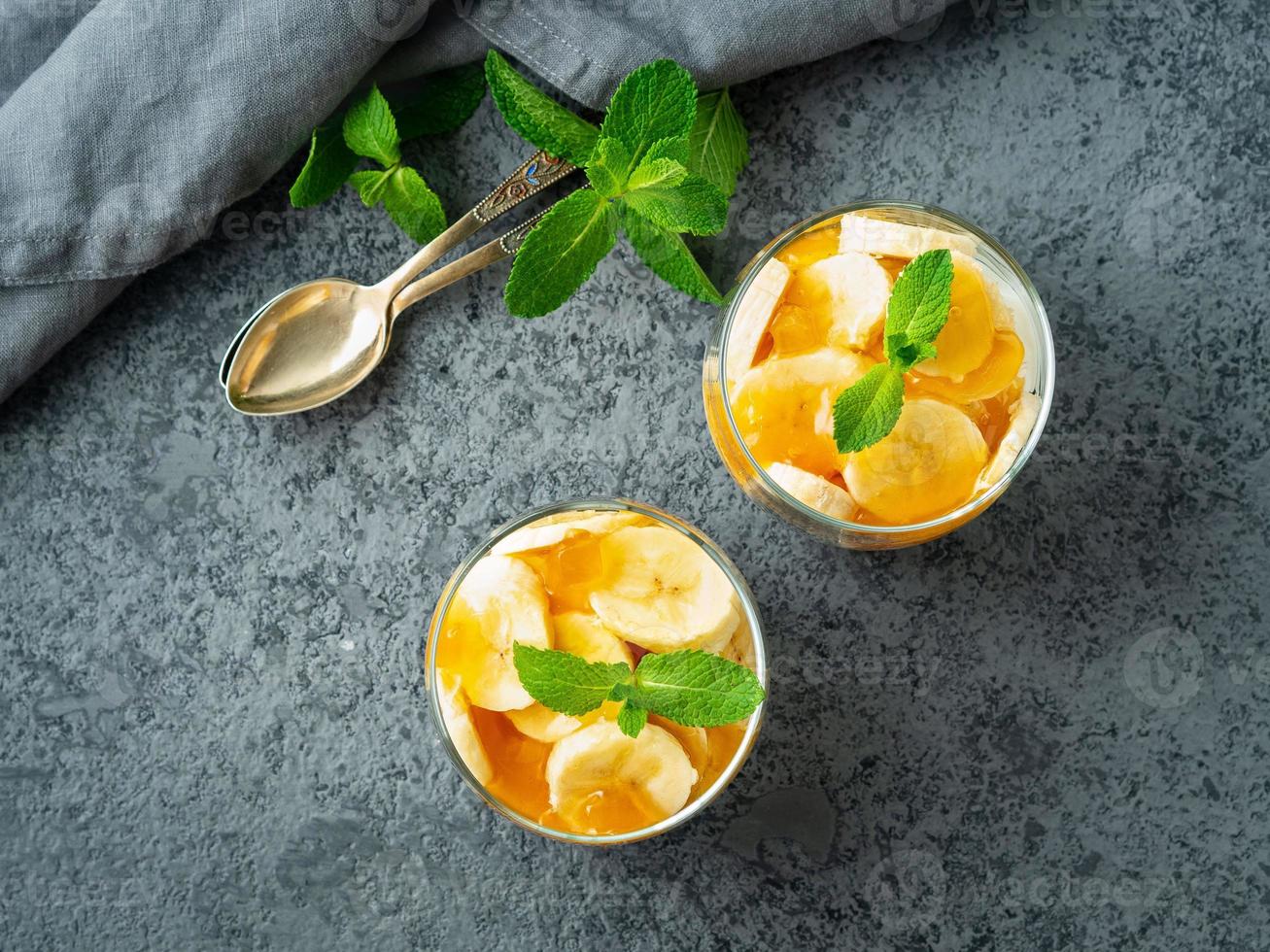 Healthy layered dessert with yogurt, banana, mango jam, cookie in glass on gray stone background, top view photo