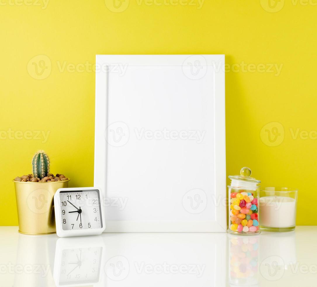 Blank white frame, clock, succulent, candy on a white table against the yellow wall. Mockup with copy space. photo