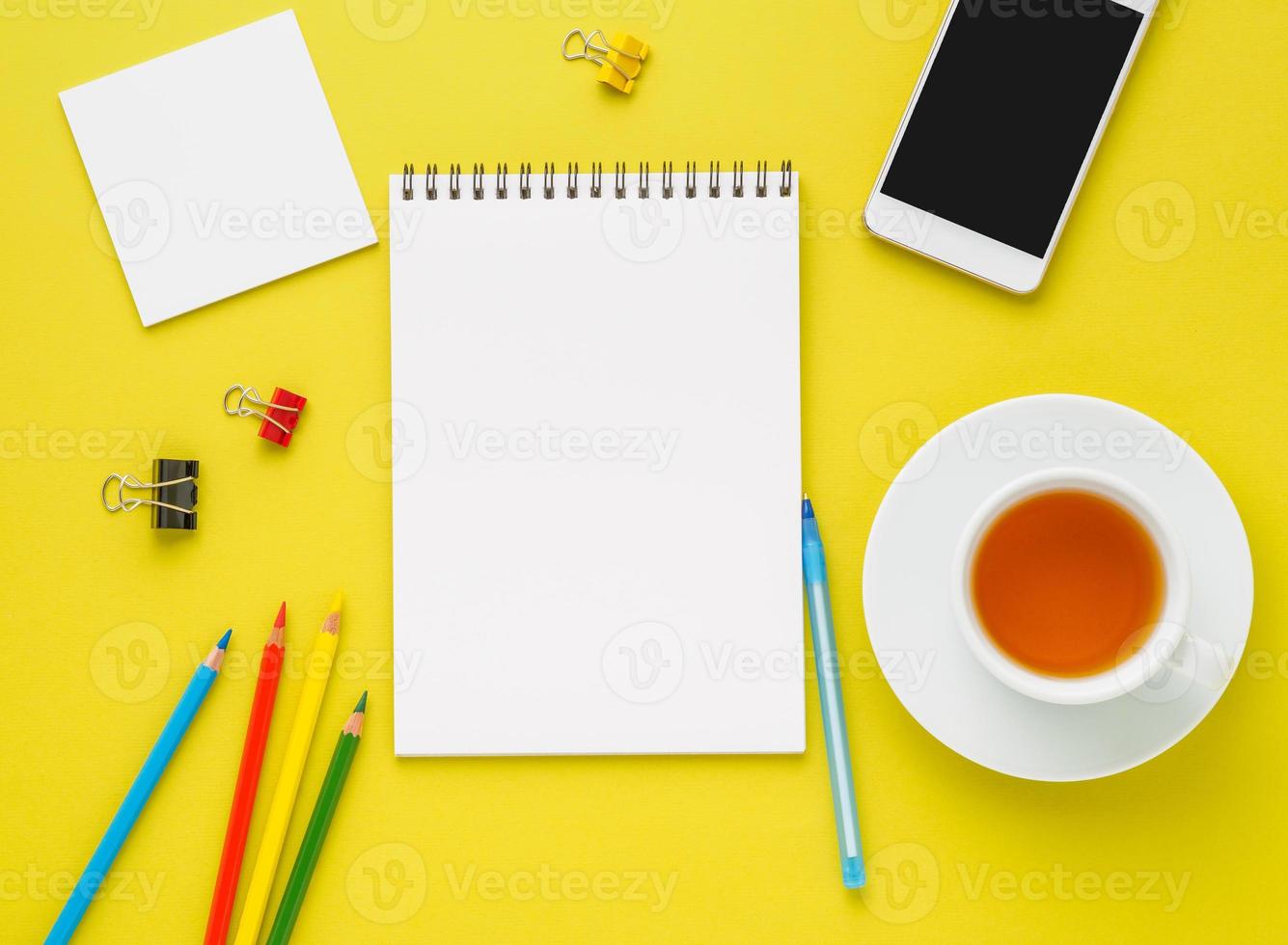 Top view of modern bright yellow office desktop with blank notepad, cup of tea and supplies. Mock up, empty space photo