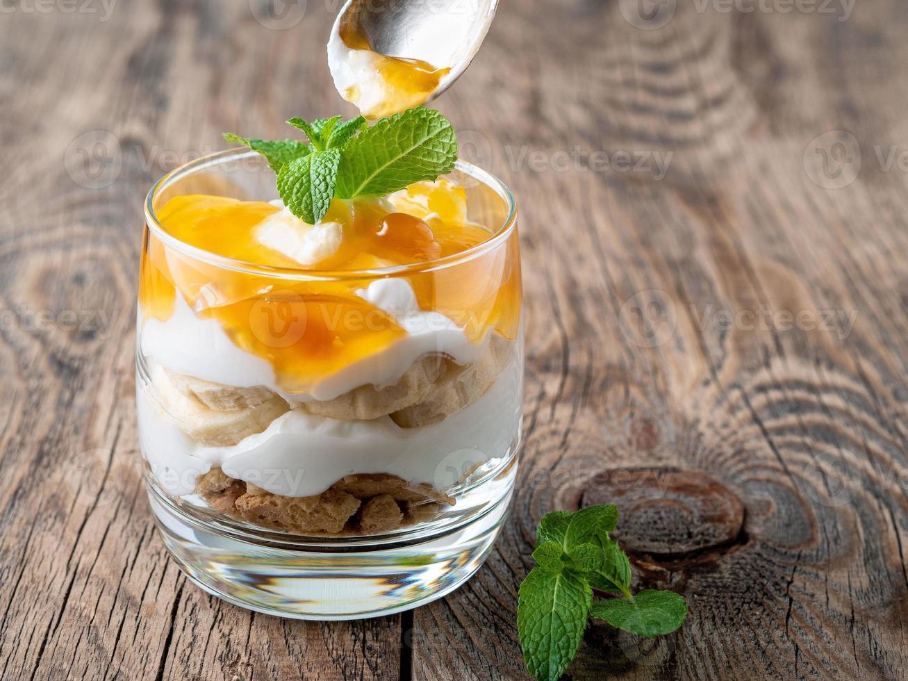 Healthy layered dessert with yogurt, banana, mango, cookie on wooden background, side view. photo