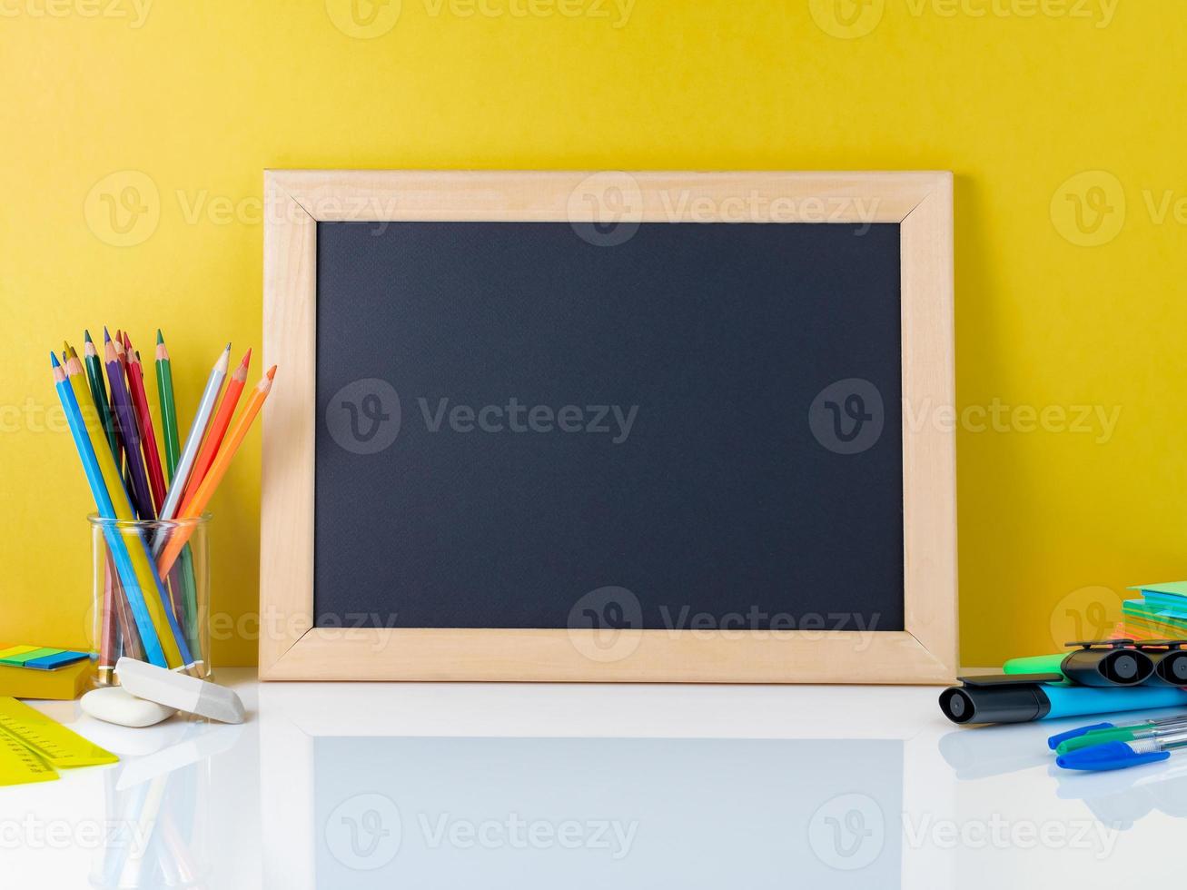 Chalkboard and school supplies on white table by the yellow wall. Side view, empty space. Back to school concept. photo