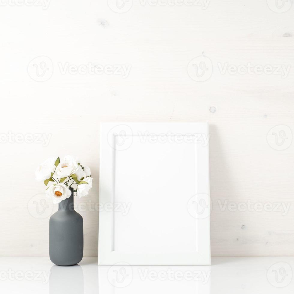 Blank white frame, flower in vaze on a white table against white wall with copy space. Mock up. photo