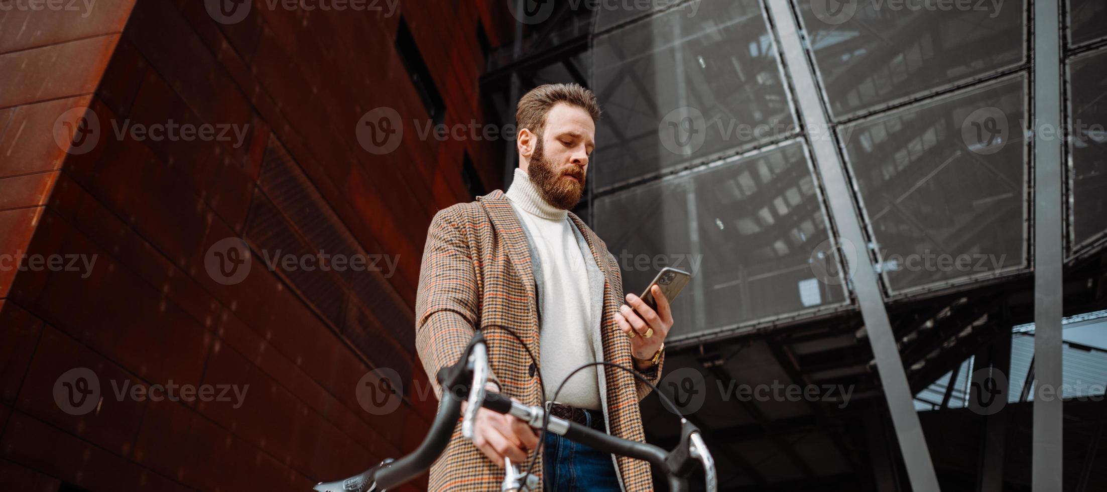 Young man with bicycle holding a smartphone front of building. Creative businessman in a modern business area. photo