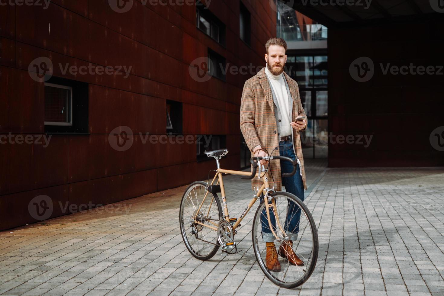 joven con bicicleta sosteniendo un teléfono inteligente frente al edificio mirando a la cámara. foto