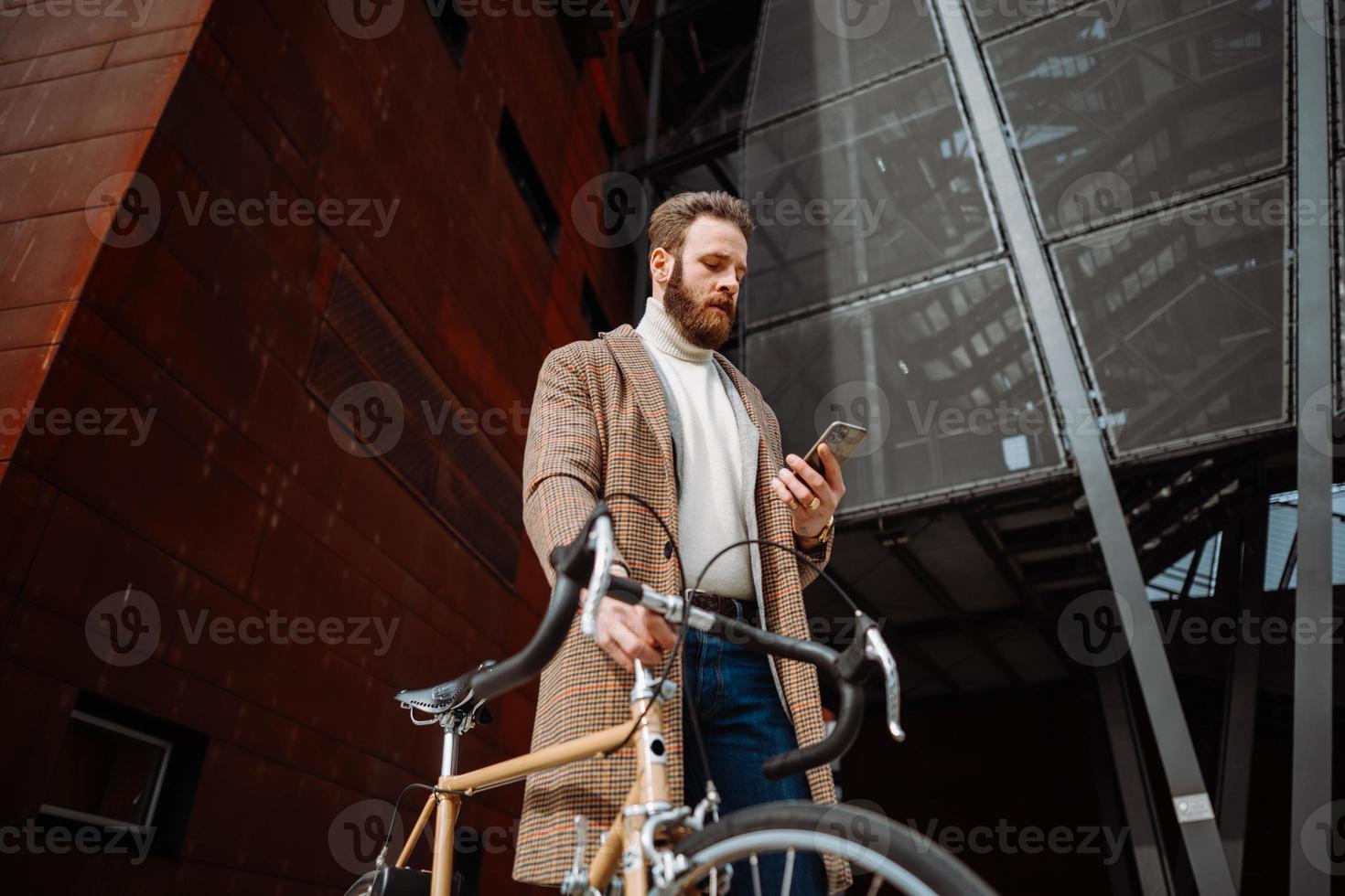 joven con bicicleta sosteniendo un teléfono inteligente frente al edificio. hombre de negocios creativo en un área de negocios moderna. foto