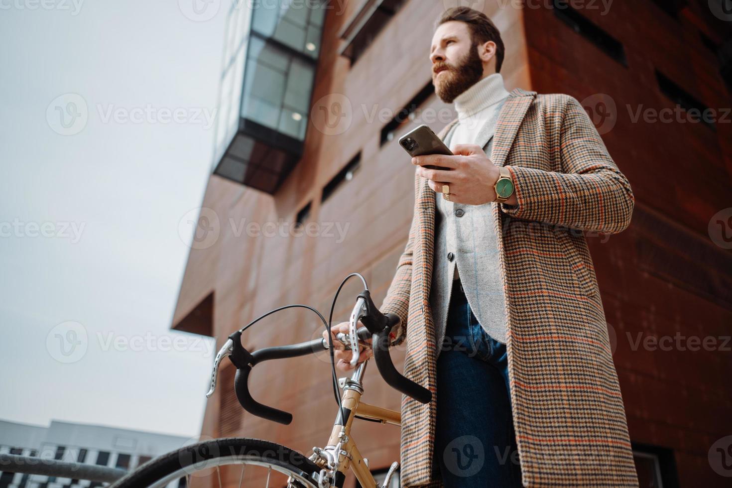 joven con bicicleta sosteniendo un teléfono inteligente frente al edificio. hombre de negocios creativo en un área de negocios moderna. foto