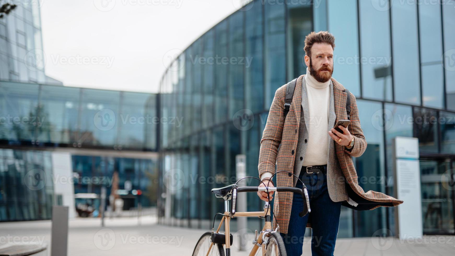 Portrait of hipster businessman with bike, using smartphone. Business centre location. Going from work. photo