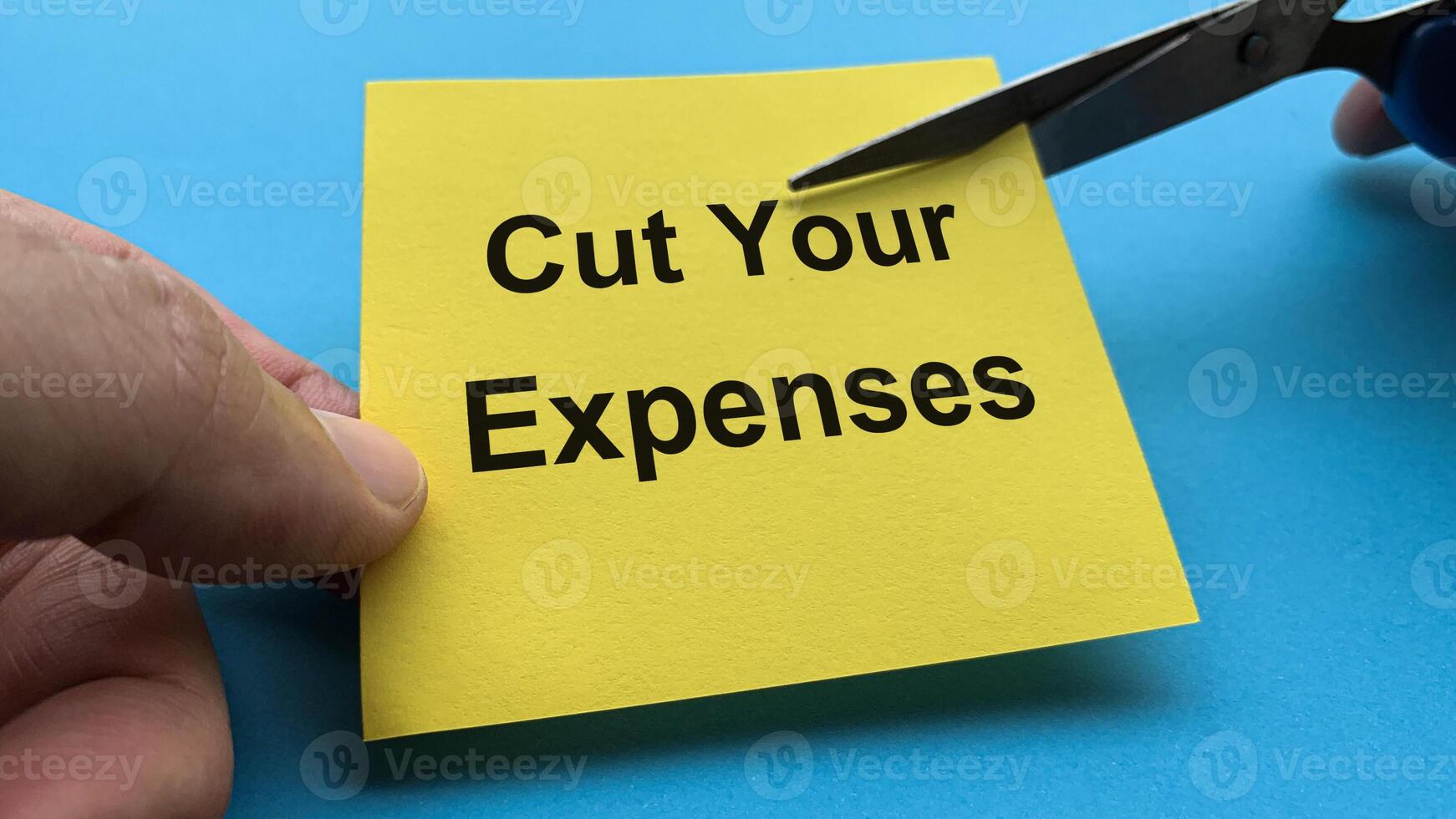 Scissors that cut yellow notepad with cut your expenses text on blue background. Spending concept photo