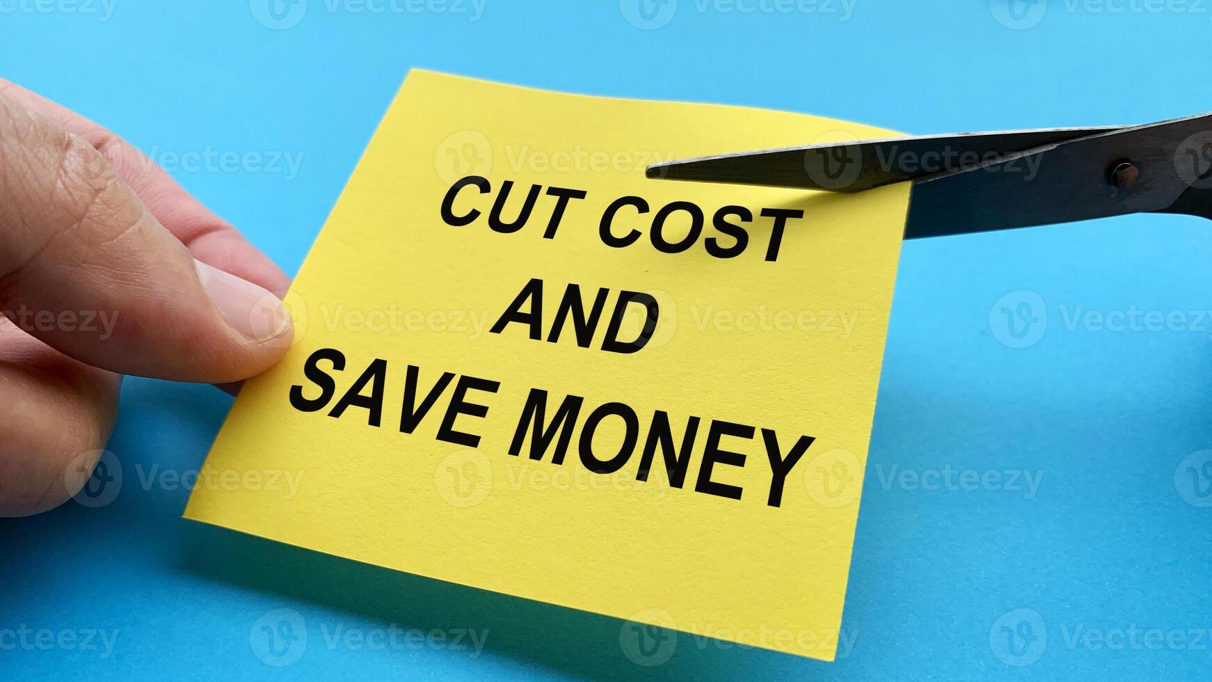 Scissors that cut yellow notepad with cut cost and save money text on blue background. Spending concept photo