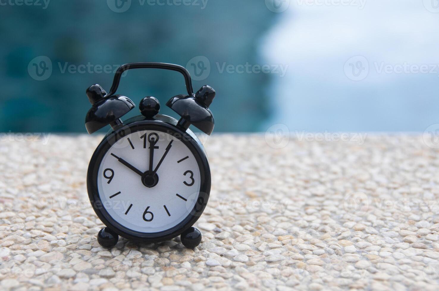 Black alarm clock on marble floor with blurred swimming pool background. The clock set at 10 o'clock. photo