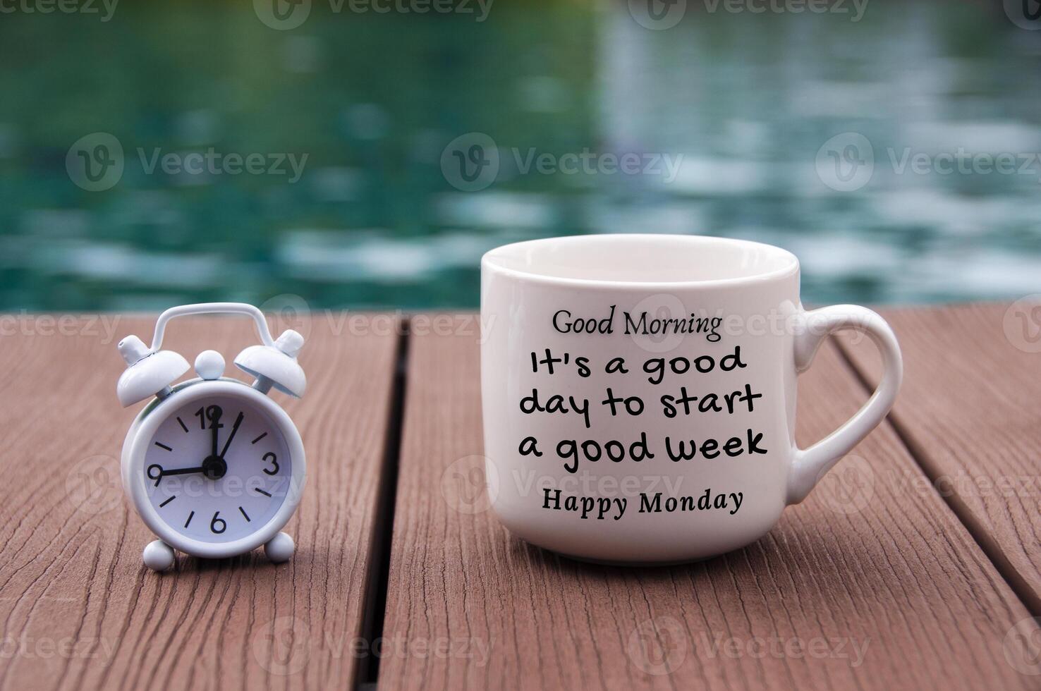 White alarm clock on wooden floor with morning quote text on coffee cup - It's a good day to start a good week. With blurred swimming pool background. photo