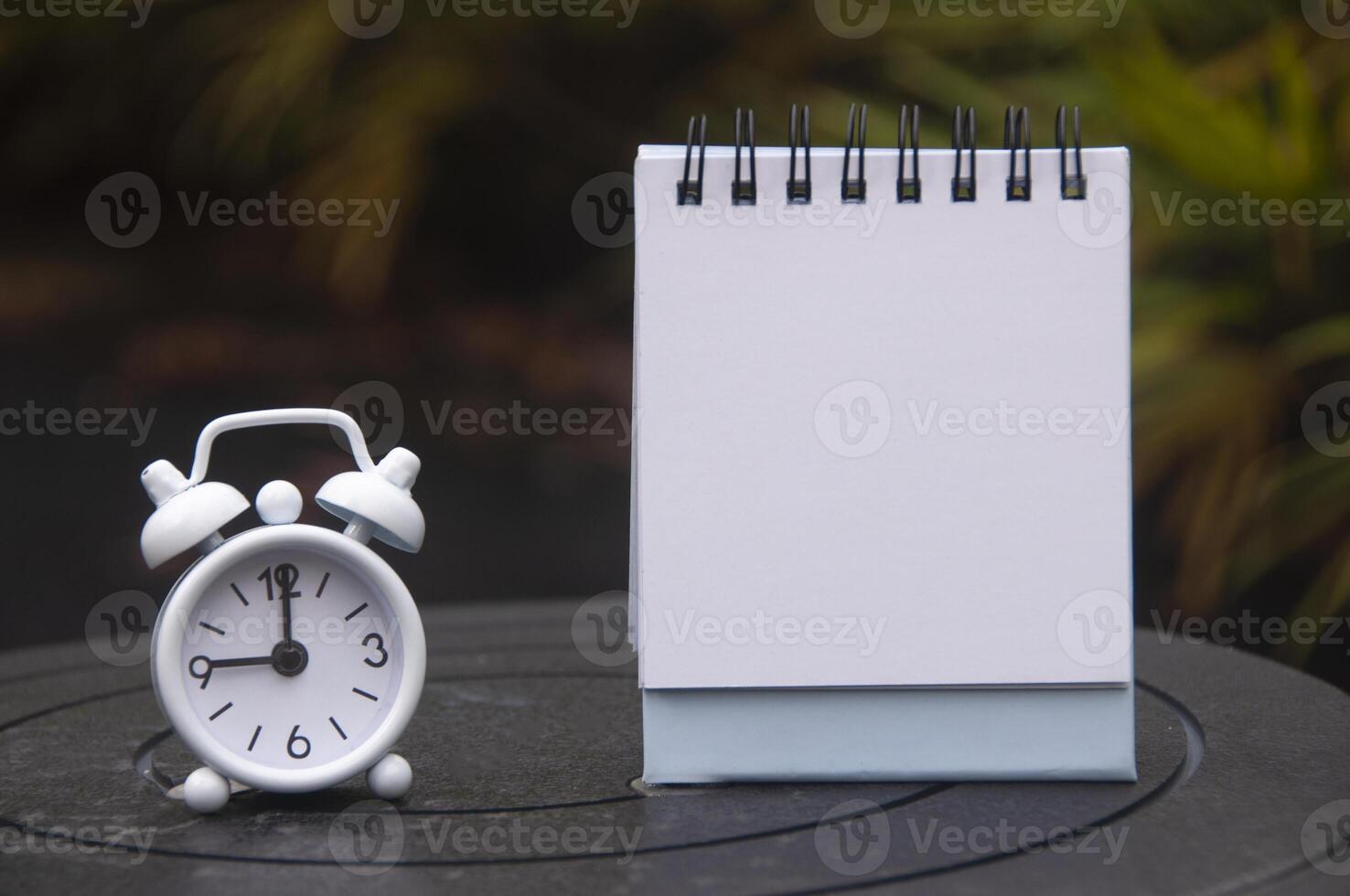 Alarm clock pointing at 9 o'clock with empty notepad. Blurred park background. Copy space concept photo