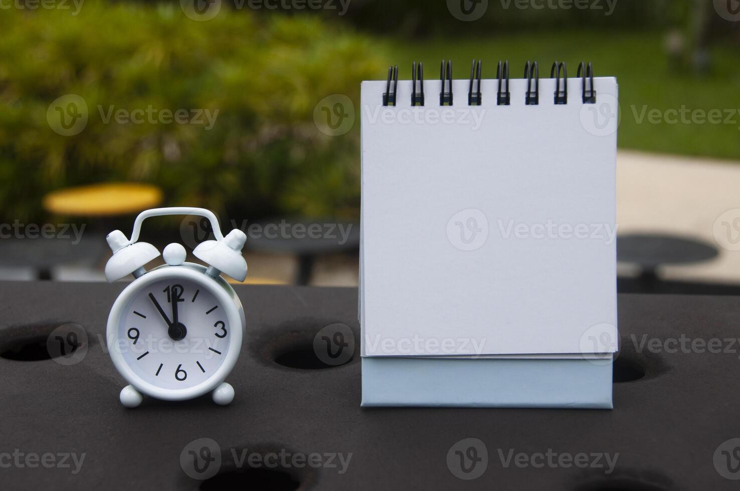 Alarm clock pointing at 11 o'clock with blank notepad. Blurred park background. Copy space concept photo