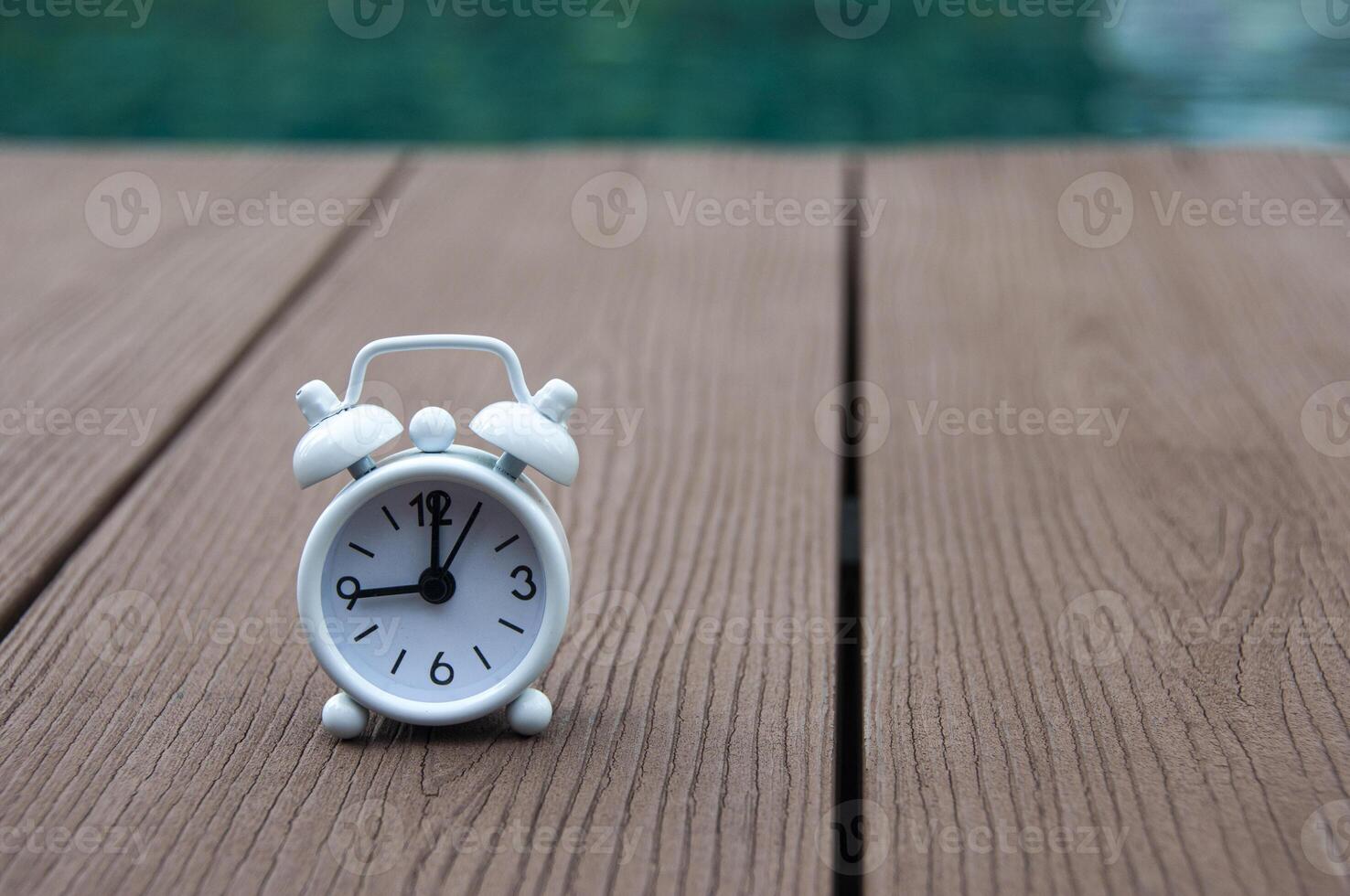 despertador blanco sobre suelo de madera con fondo de piscina borroso. el reloj puesto a las 9 en punto. foto