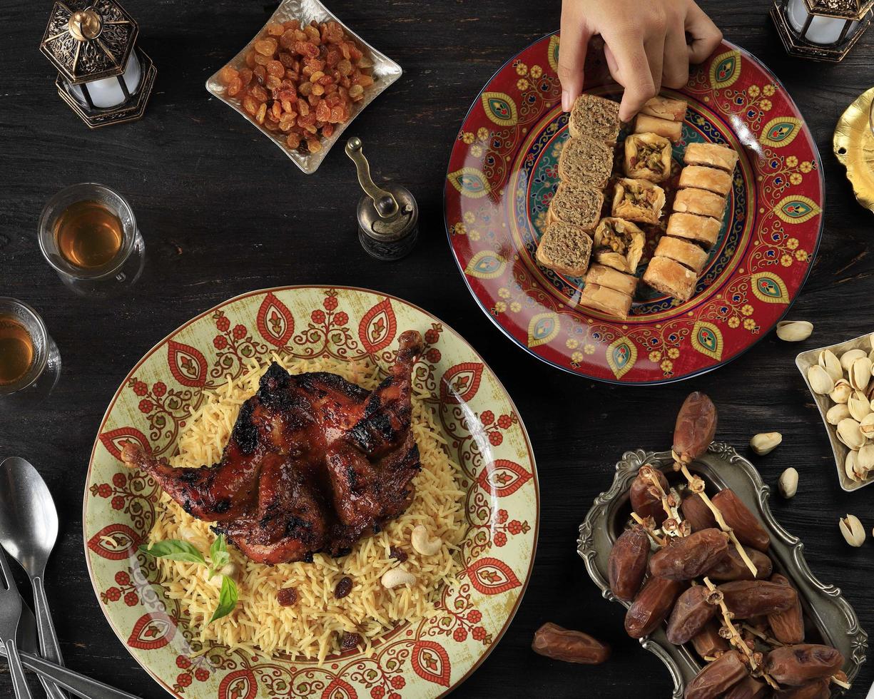 mano femenina elige baklava turco en el plato, mesa para el saludo de ramazán con dátiles, arroz árabe, pollo asado, nueces y té. foto