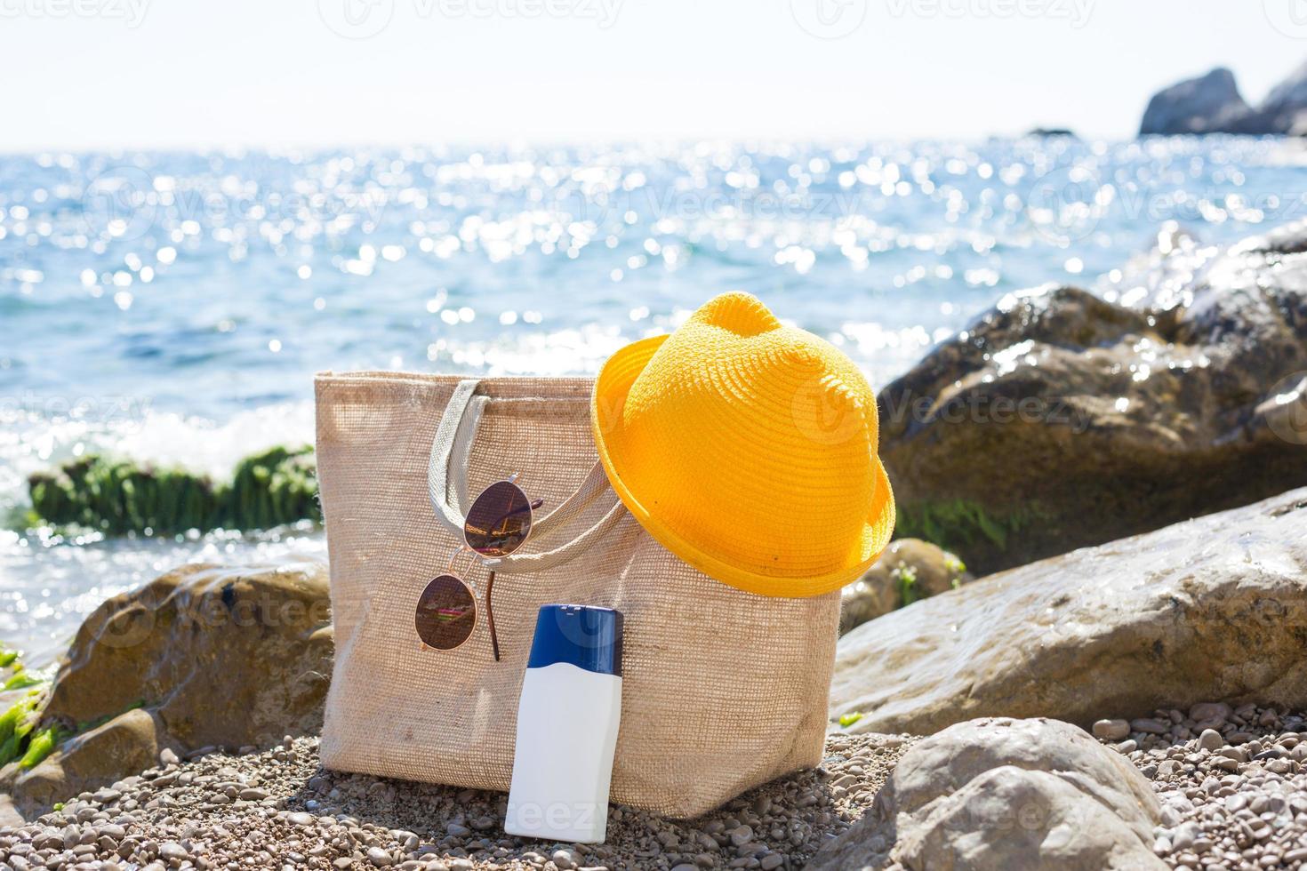 Beach bag with a white tube of sunscreen on a pebble beach near the sea. Travel, beach holiday at the resort, glasses, yellow hat. Protection of the skin from UV rays, suntan oil. Copy space, mock up. photo
