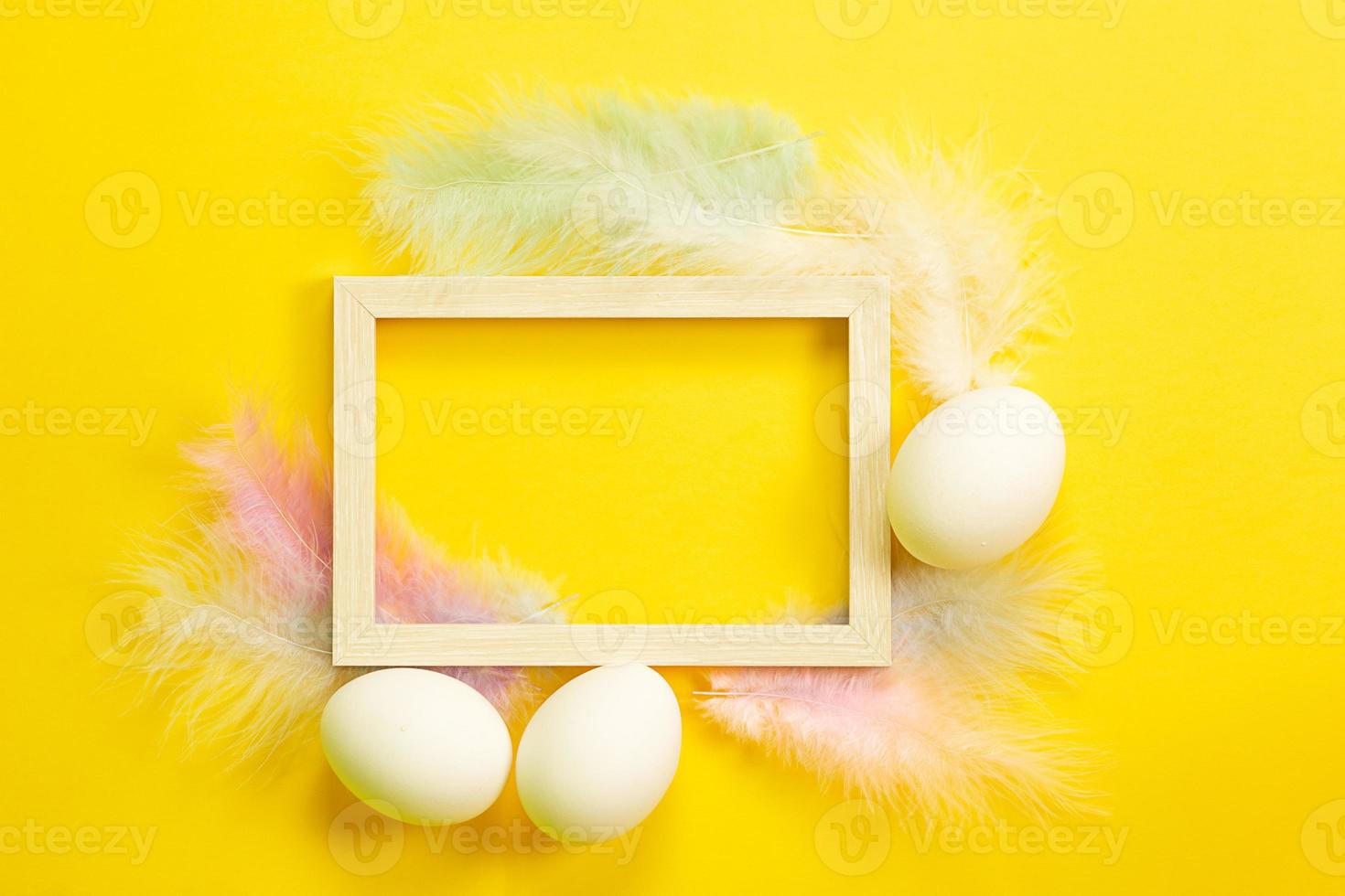 Easter white frame on a bright yellow background of chicken eggs and colored delicate feathers. Spring, religious holiday, Easter decoration, greeting, copy space, mock up photo