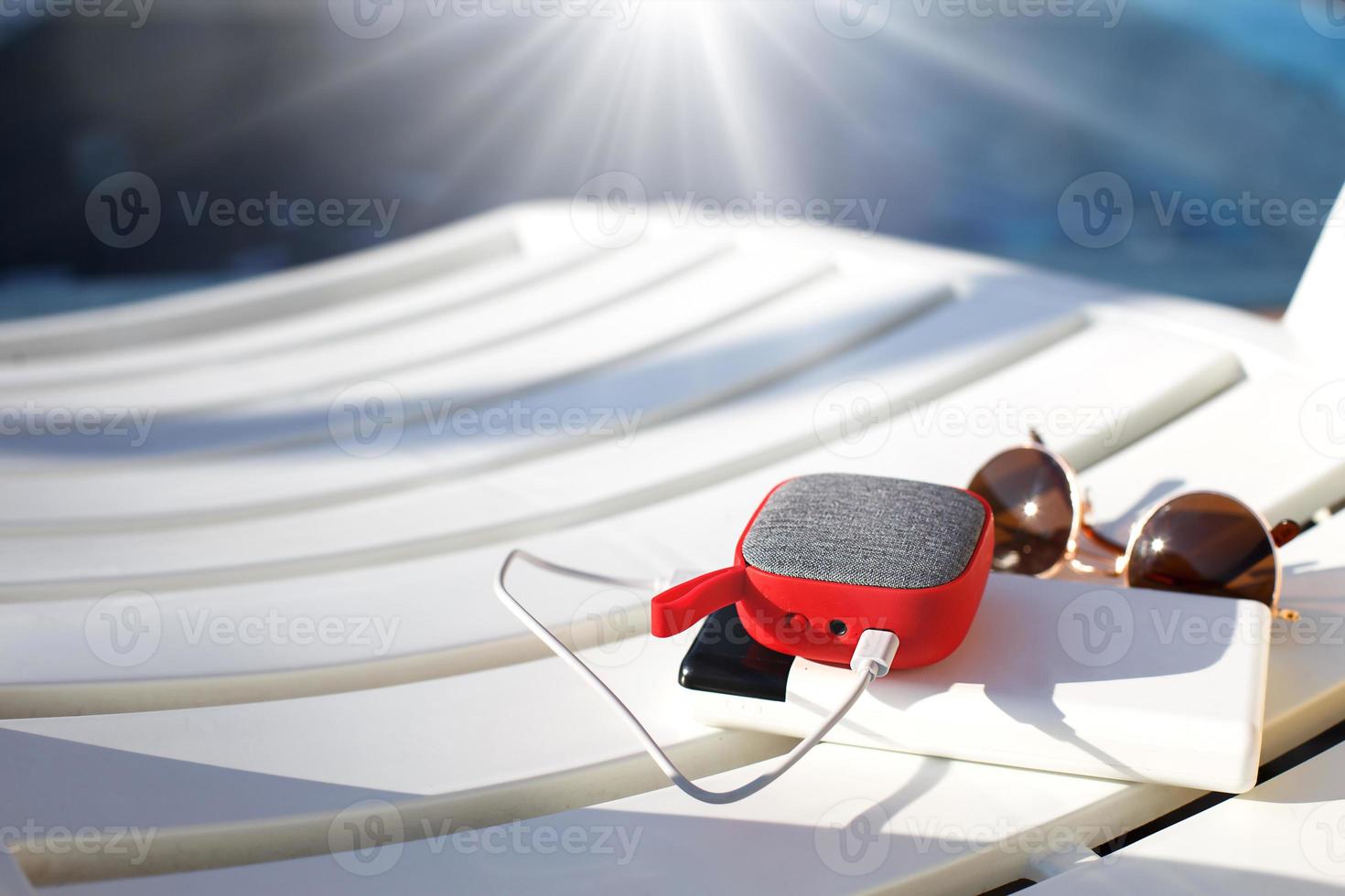 Red music portable speaker is charged from the power bank via usb on a deck chair near the pool. Concept is always in touch, travel gadgets, external battery for a smartphone. Place for text. Flatlay photo