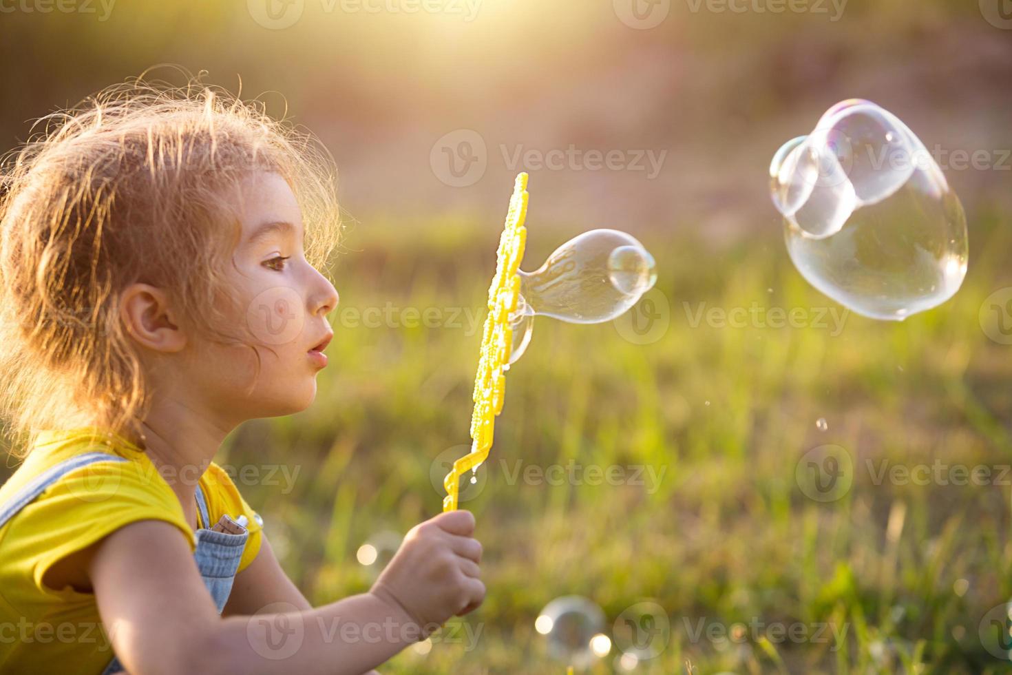 A girl in a denim jumpsuit blows soap bubbles in the summer in a field at sunset. International Children's Day, happy child, outdoor activities. Summer background. Healthy and eco-friendly lifestyle photo