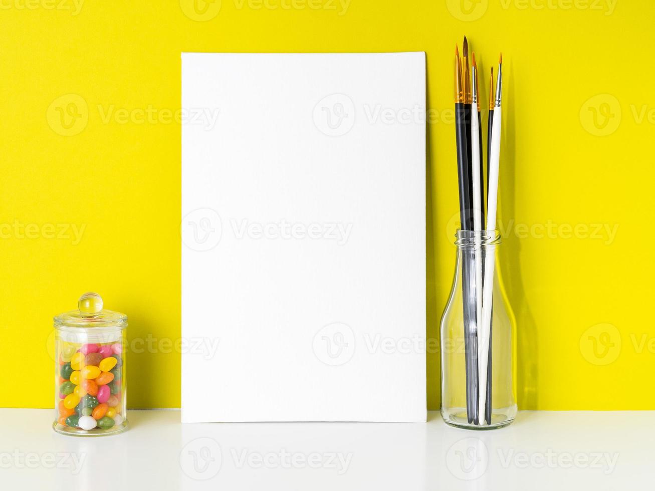 Mockup with clean white canvas, candy in jar, brushes on bright yellow background. Concept for creativity, drawing. photo