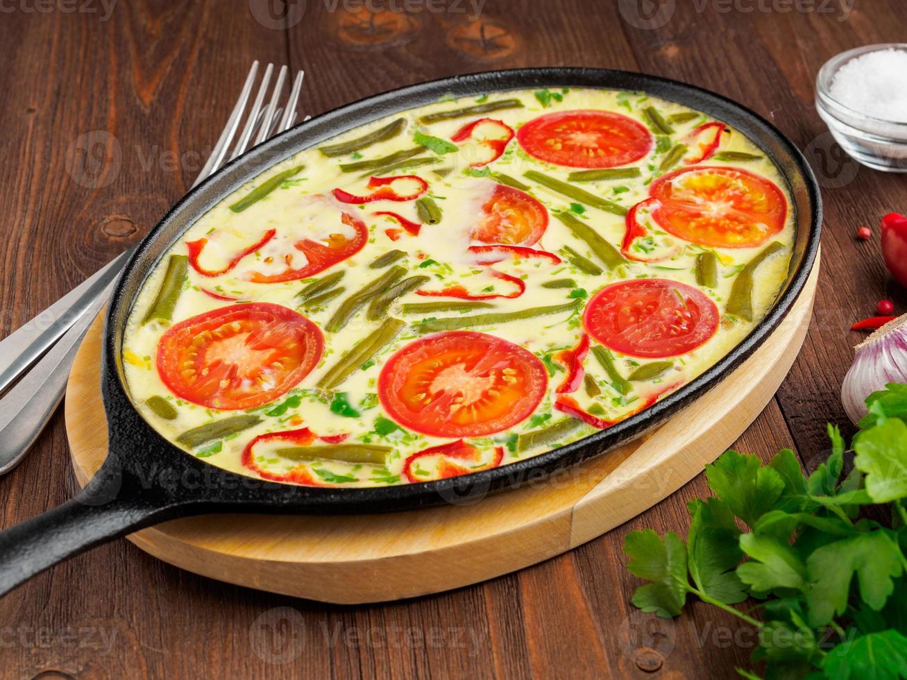 Oval cast iron pan with delicious hot omelette with tomatoes and capsicum. Brown wood texture background, close-up, side view. photo