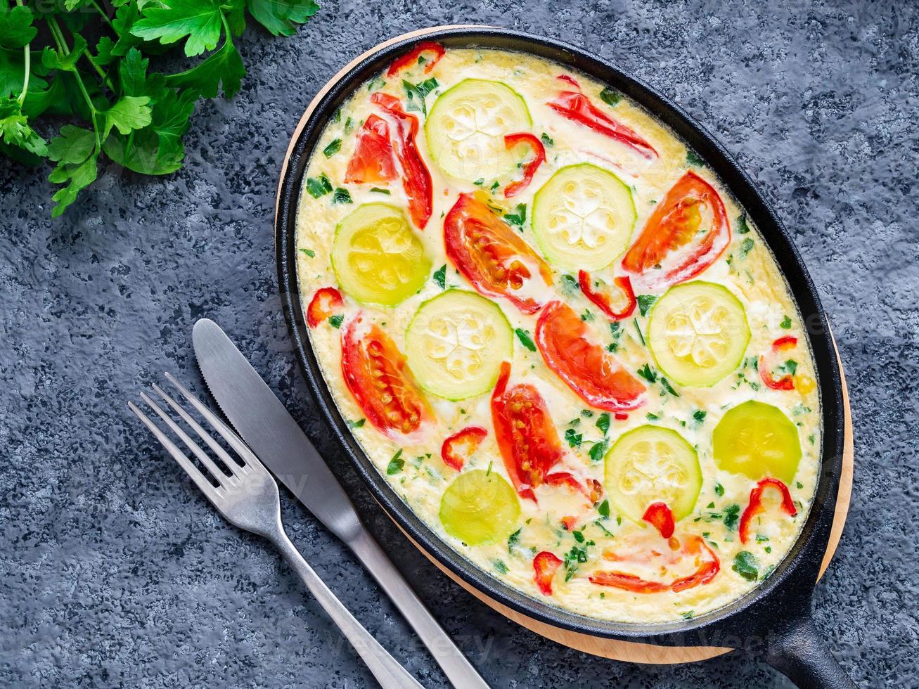 sartén con frittata italiana tradicional sobre mesa de piedra gris oscuro, tenedores, perejil, pimienta, tomates. huevos revueltos foto