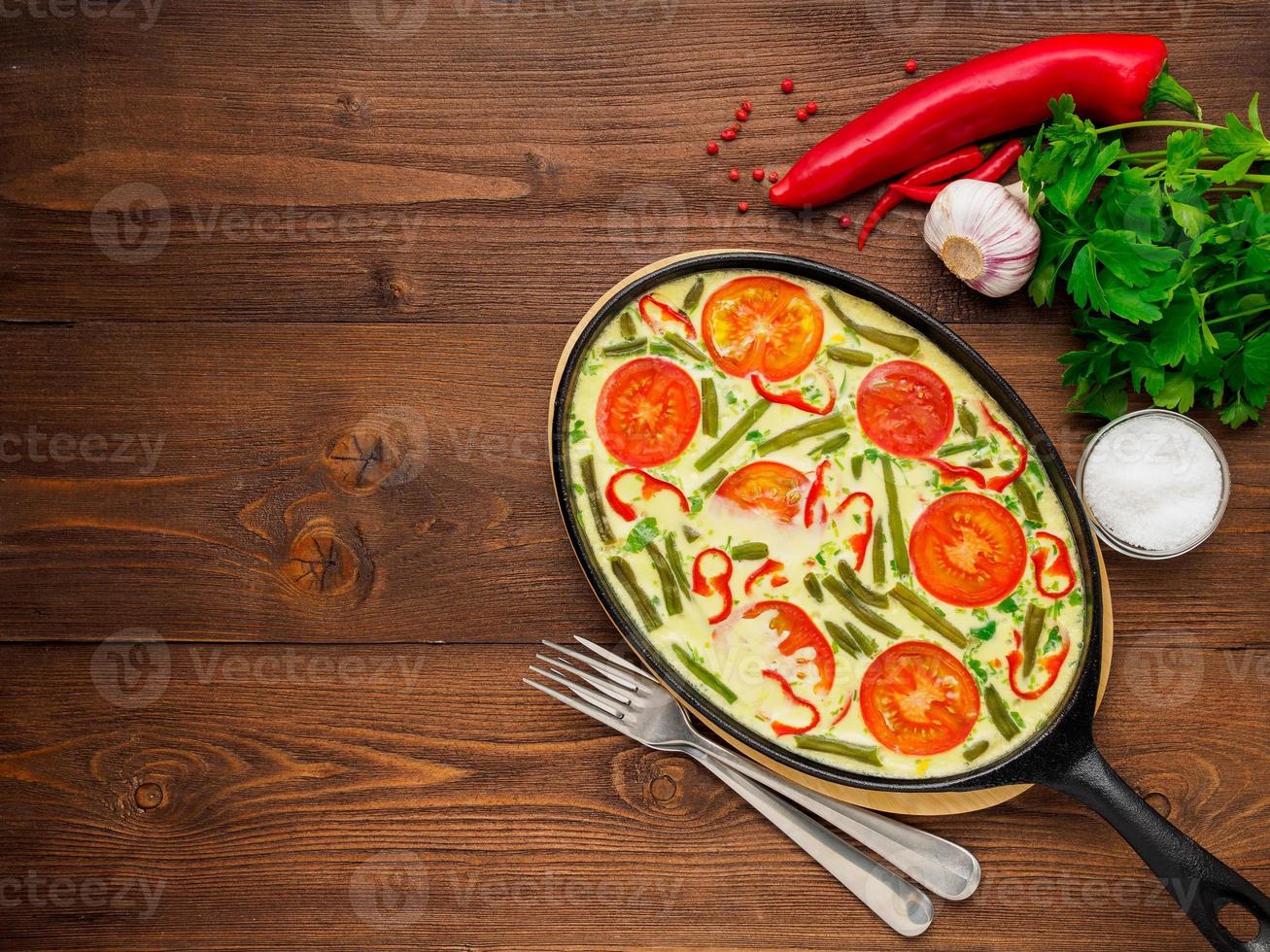 Pan with traditional Italian Frittata on the side of a brown wooden table, forks, parsley, salt, pepper, tomatoes. Top view, empty space for text photo