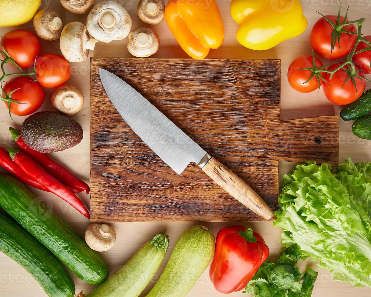Vegetables around wooden cutting board with knife on kitchen table. Top view. photo