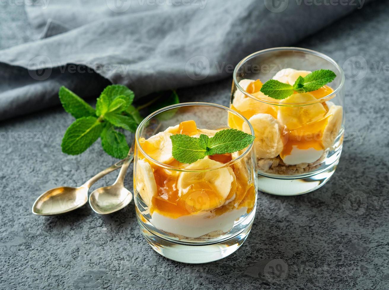 Healthy layered dessert with yogurt, banana, mango jam, cookie in glass on gray stone background, side view photo