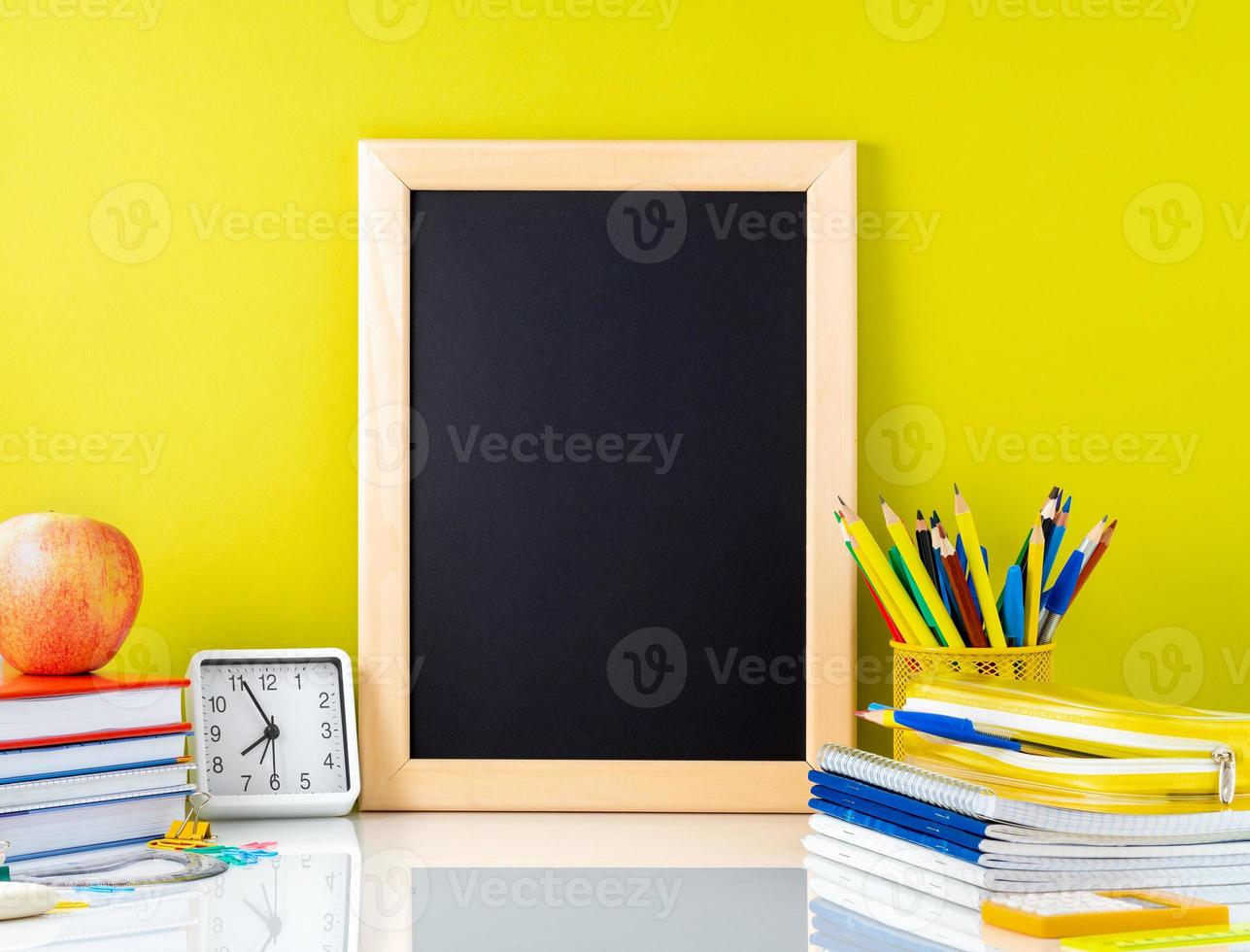 Chalkboard, apple and school supplies on white table by the yellow wall. Side view, empty space. Back to school concept. photo