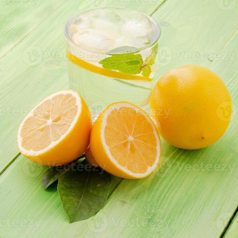 Lemonade in a glass, a lemon half, fresh leaves on the green table. A refreshing cold drink of water with ice, mint and slices of lemon, side view photo