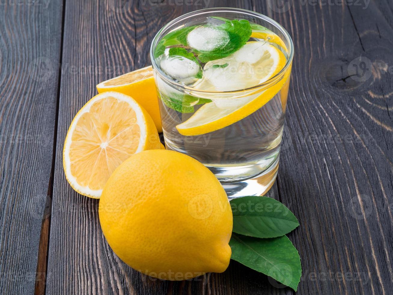 Lemonade in a glass, a lemon half, fresh leaves on dark brown wood table. A refreshing cold drink of water with ice, mint and slices of lemon, side view photo