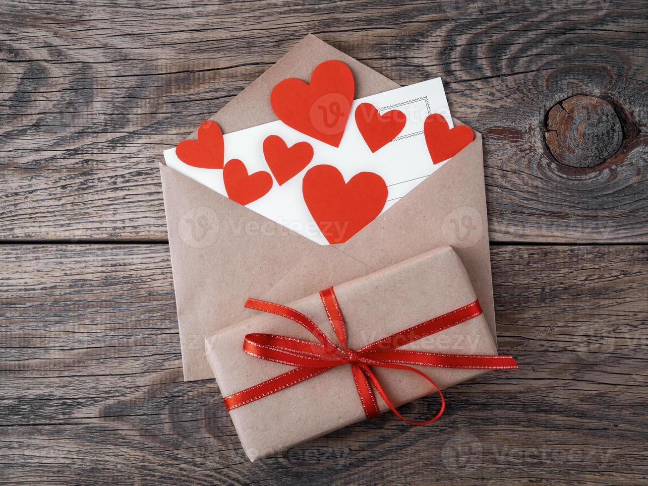 card and red hearts in open envelope from brown Kraft paper. Gift box with red ribbon on wooden aged vintage background. Greetings with Valentine's day photo