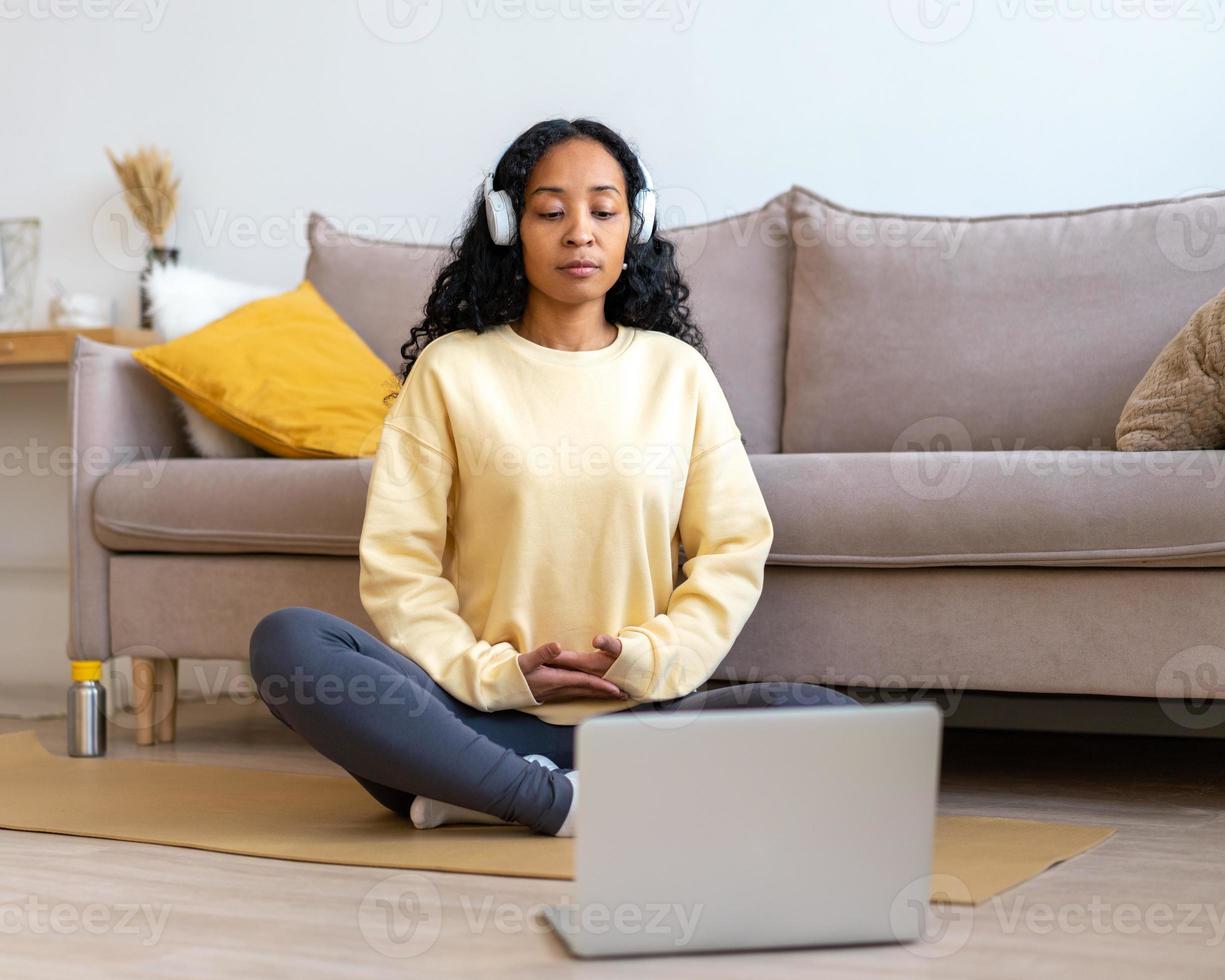 mujer afroamericana escuchando música con auriculares mientras medita con una laptop en casa foto