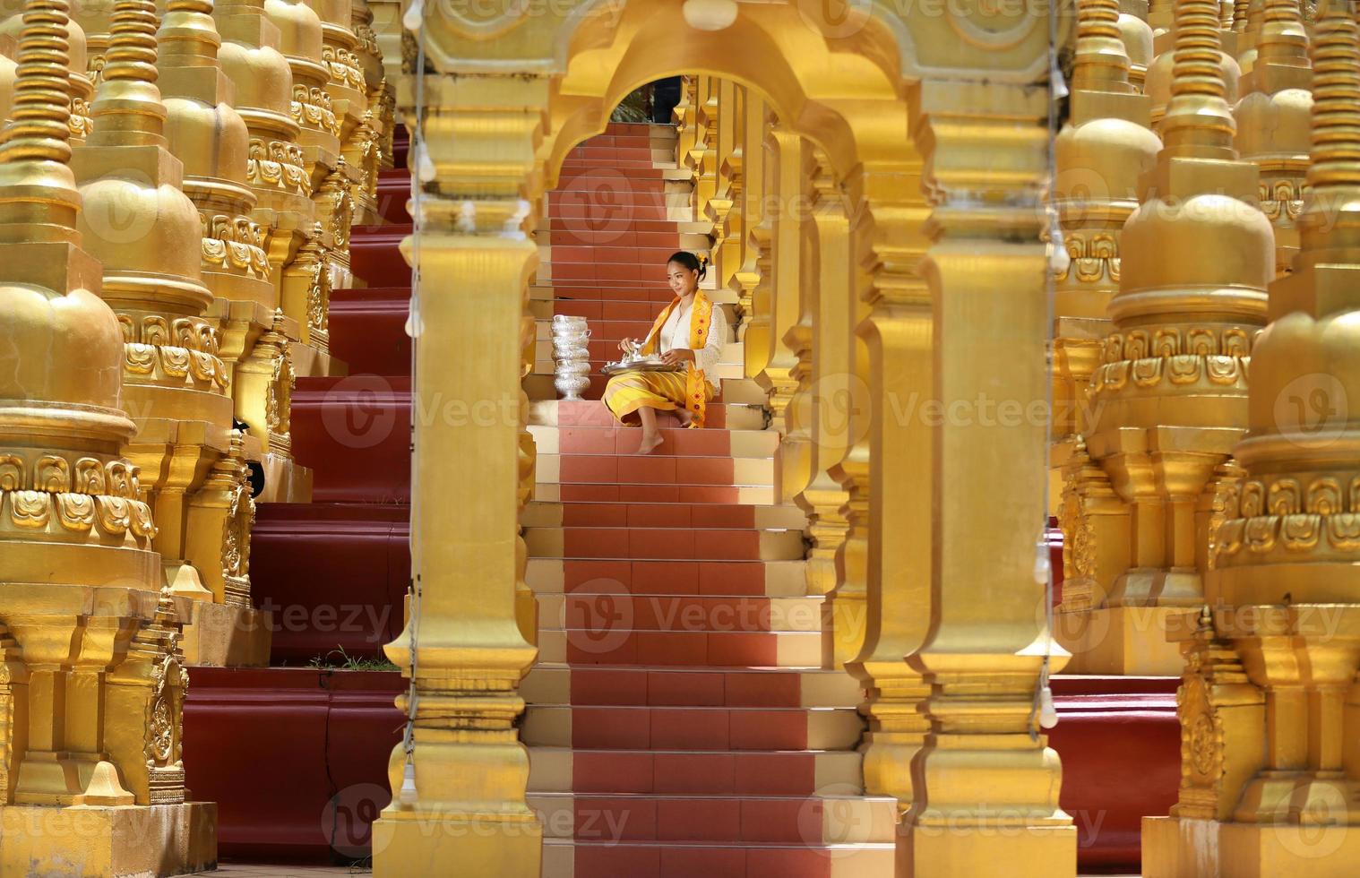 joven asiática con traje tradicional birmano sosteniendo un tazón de arroz a mano en la pagoda dorada en el templo de myanmar. mujeres de myanmar sosteniendo flores con vestido tradicional birmano visitando un templo budista foto