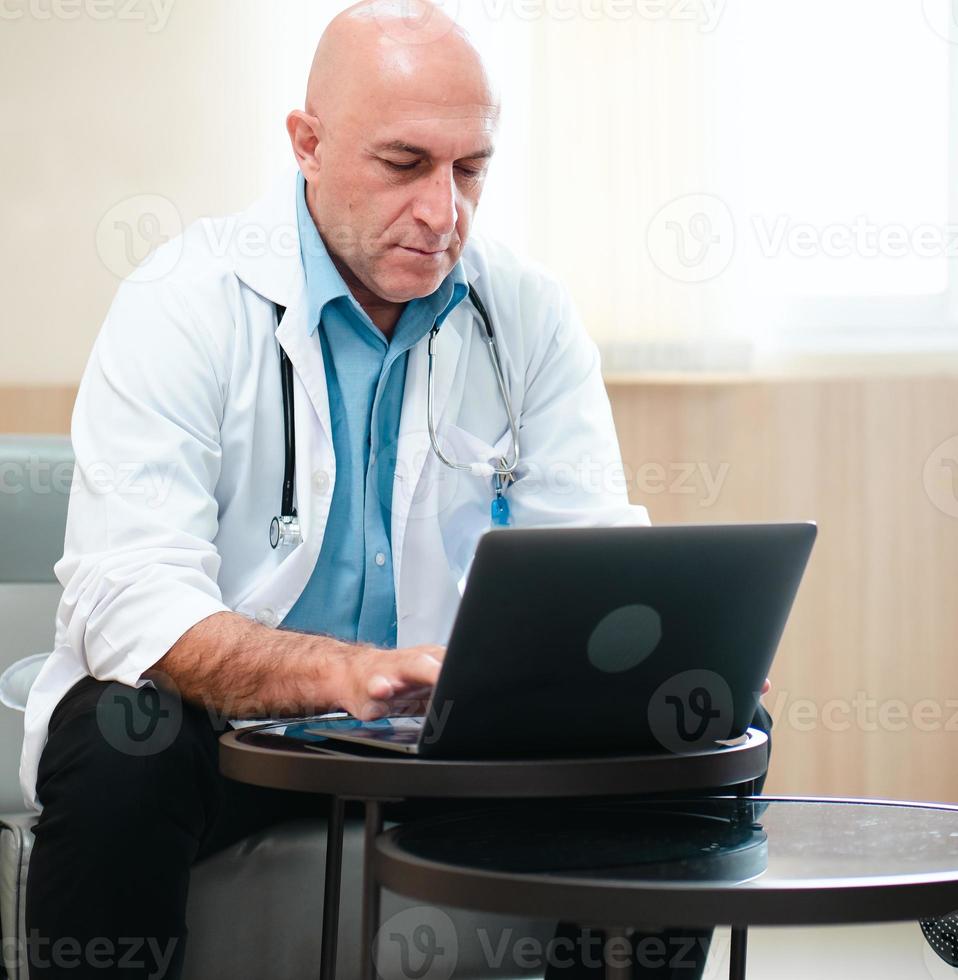 Male doctor is using notebook computer in hospital living room. Professional medical person in white coat and stethoscope sits relaxing looking at computer screen for diagnosis, healthcare information photo