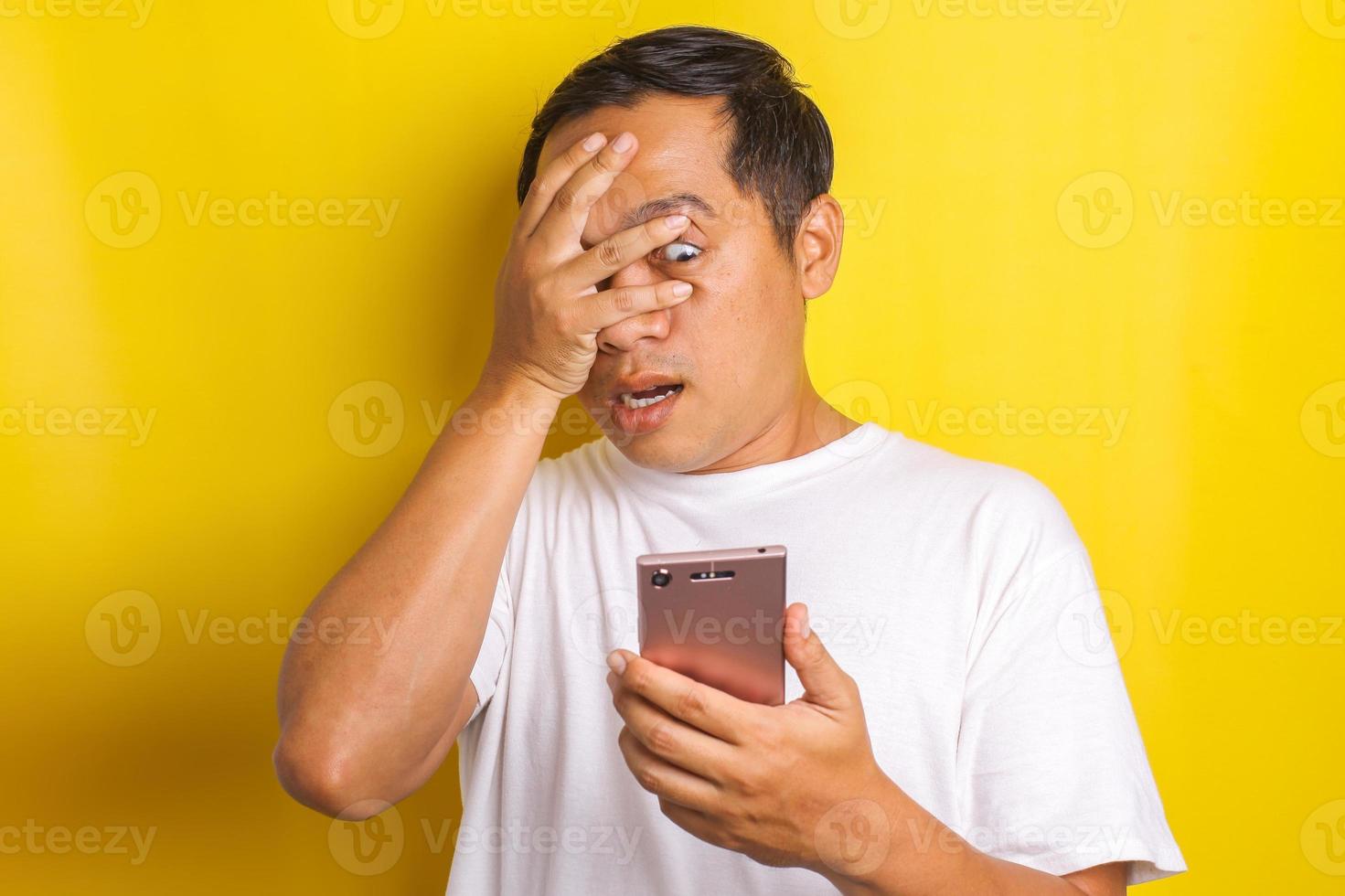 Close-up portrait of shocked, scared asian man closing one eye while looking at cell phone isolated on yellow background photo