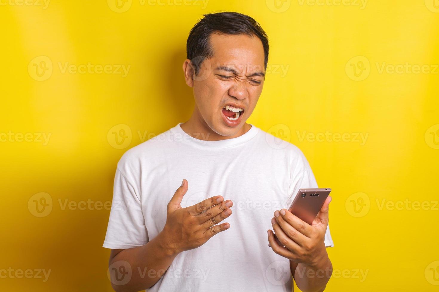 Close-up portrait of asian man with angry expression, annoyed, shouting looking at cell phone isolated on yellow background photo