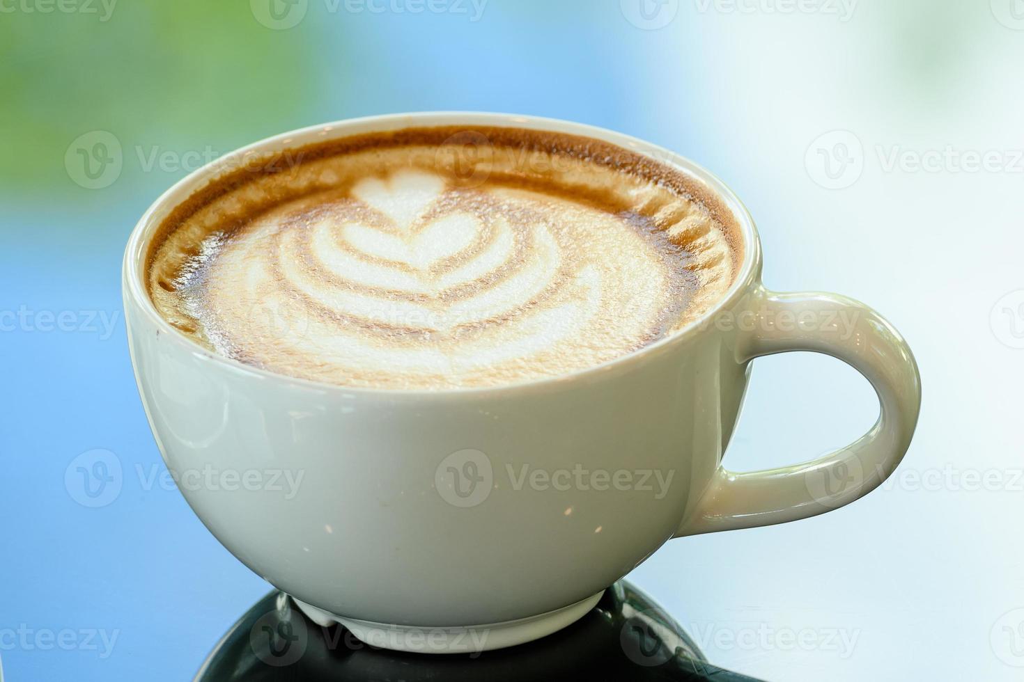 Latte art coffee cup on glass table. Blurred leaf reflection background. photo