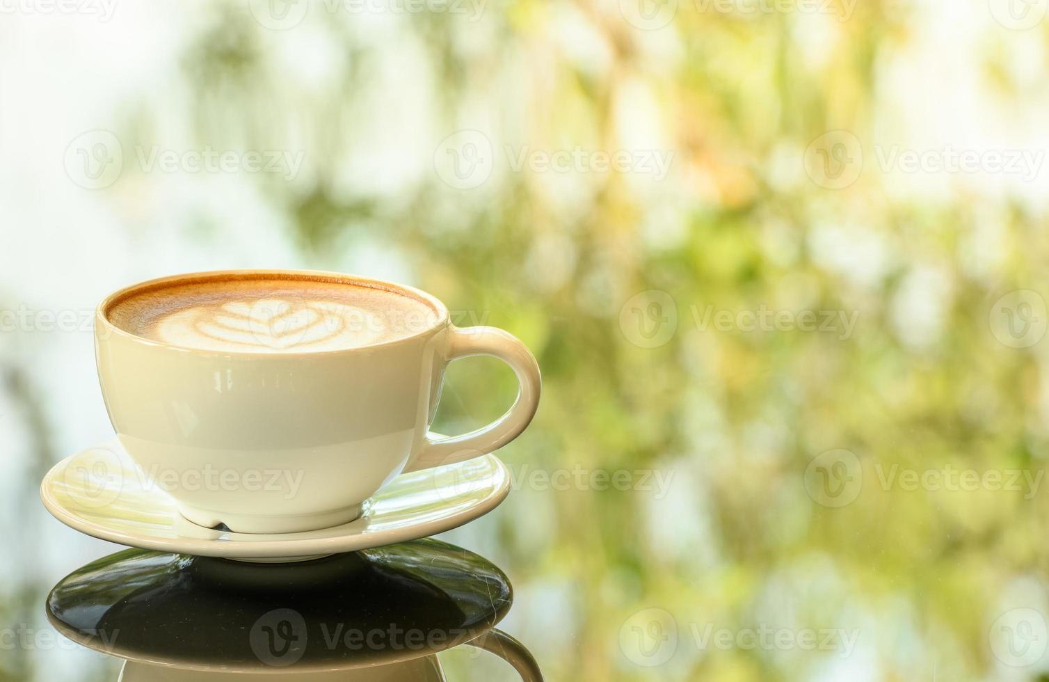 Latte art coffee cup on glass table. Blurred leaf reflection background. photo