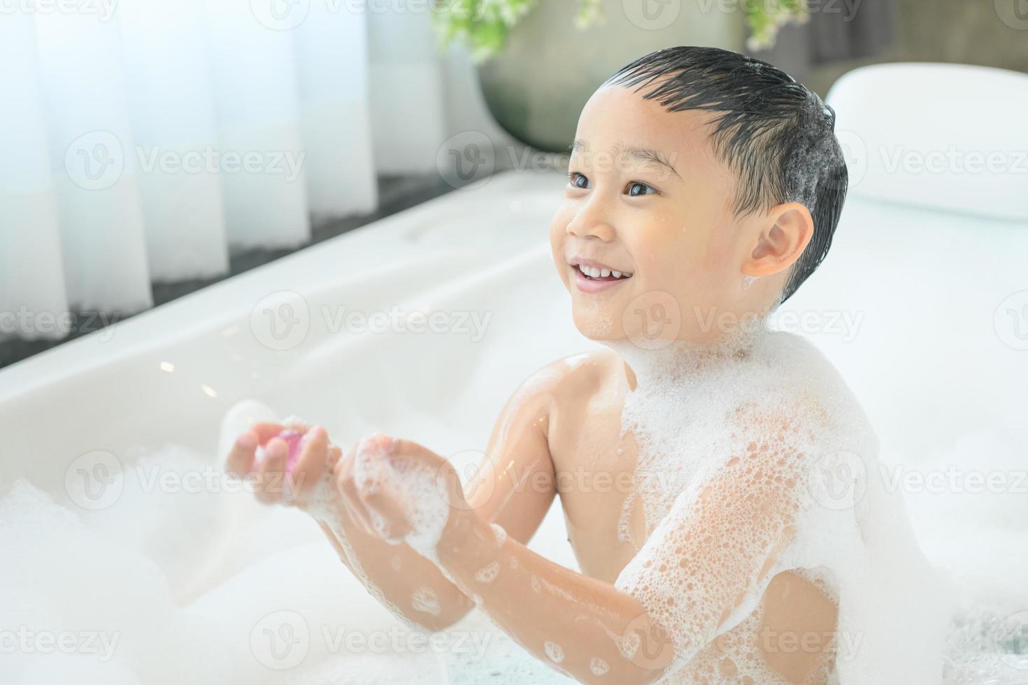 smiling happy asian child boy is playing with white foam in tub bath at home. photo